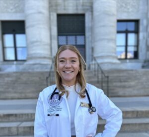 A smiling female doctor in a white coat with a stethoscope outdoors.